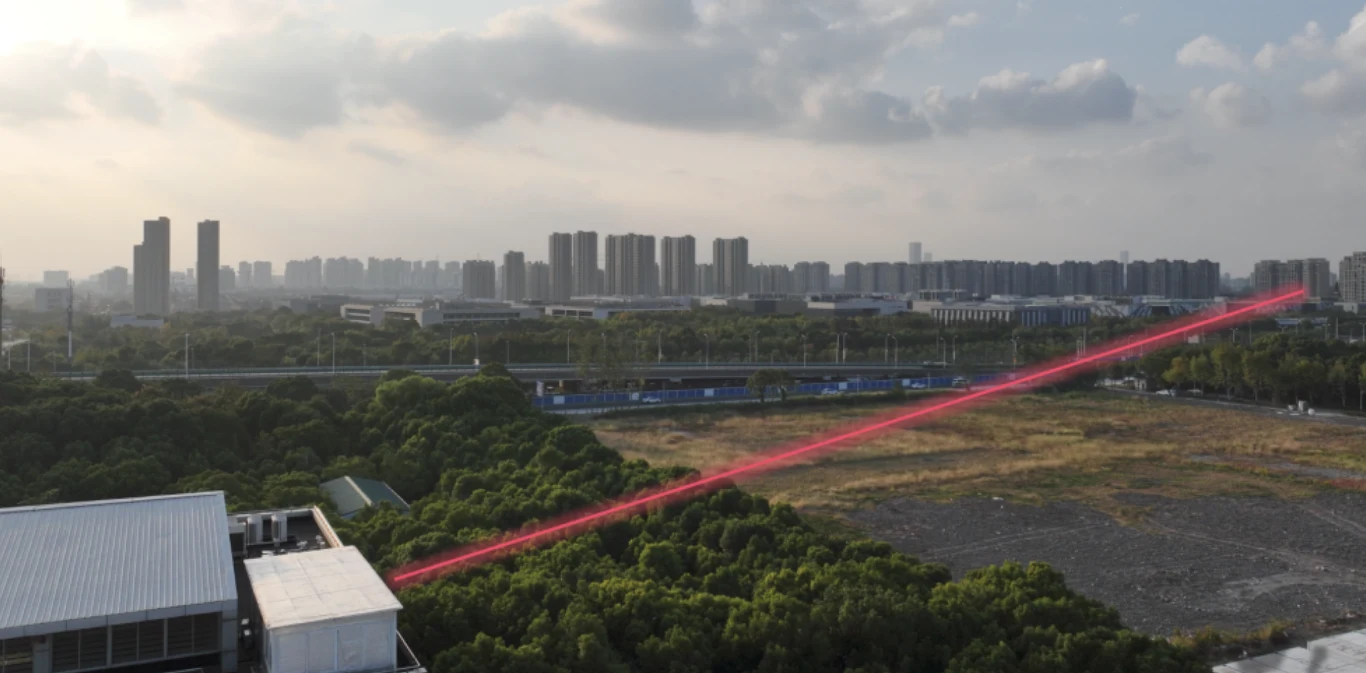 System zdalnego obrazowania emituje osiem wiązek lasera podczerwonego w kierunku celu znajdującego się w budynku oddalonym o 1,36 km Widok miejskiego krajobrazu z licznymi blokami mieszkalnymi i rozległym terenem zielonym, przez środek zdjęcia przebiega intensywnie widoczna, czerwona wiązka światła laserowego.