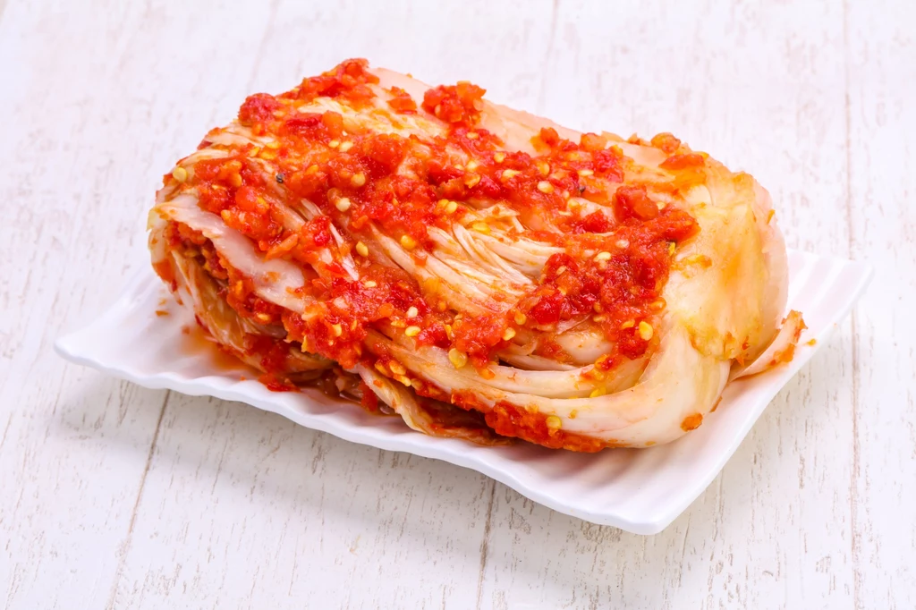 KImchi to przysmak kojarzony z kuchnią koreańską Porcja kimchi, czyli pikantnej koreańskiej fermentowanej kapusty pekińskiej z dodatkiem przypraw i chili, ułożona na białym talerzu na jasnym, drewnianym tle.