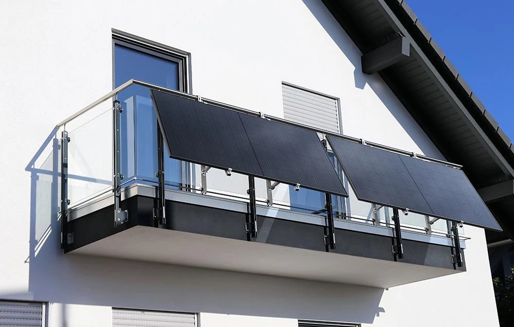 STREAM udowadnia, że ekologiczne wybory są możliwe także w bloku Nowoczesny balkon z przeszkloną balustradą, na którym zamontowano panele fotowoltaiczne nachylone pod kątem, umieszczony na białej elewacji budynku pod dachem dwuspadowym; w tle widoczne okna i rolety zewnętrzne.