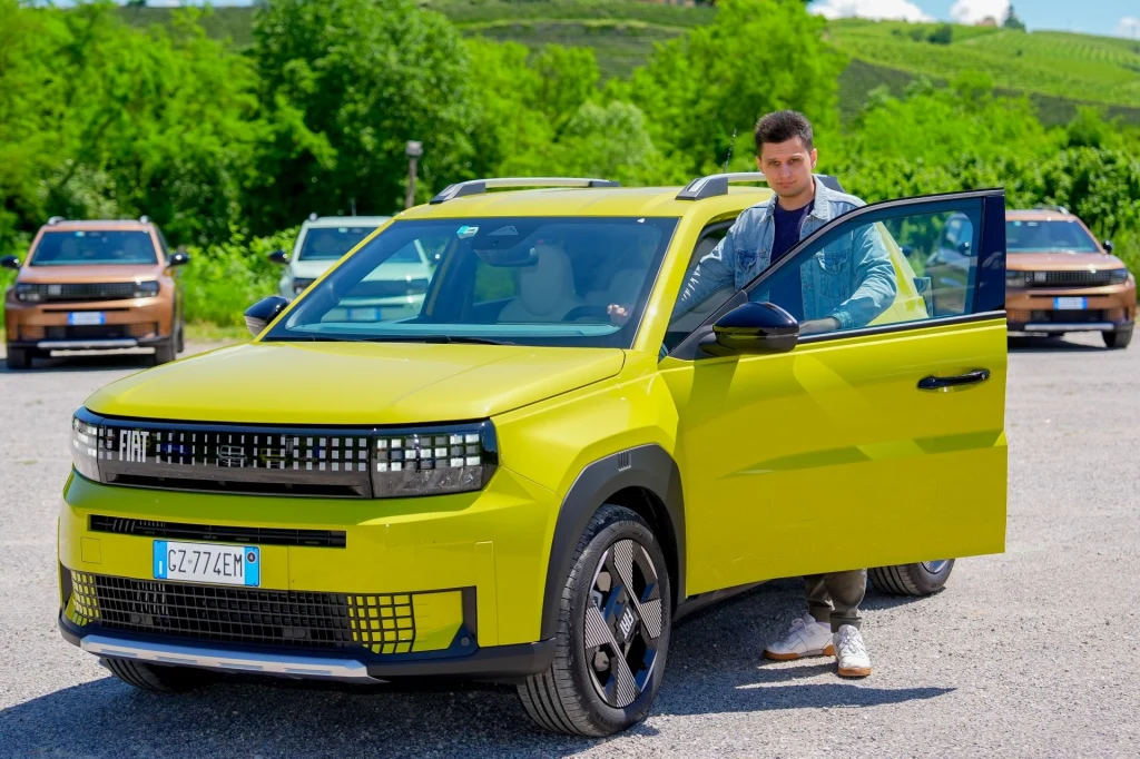 Jaki jest najlepszy tani samochód elektryczny dostępny w Polsce? Mężczyzna stojący przy otwartych drzwiach nowoczesnego żółtego samochodu marki Fiat, w tle kilka innych aut tej samej marki na parkingu na tle zieleni i wzgórz.