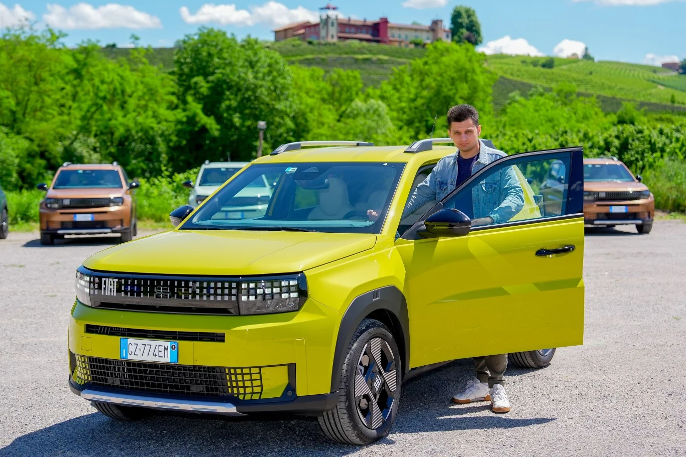 Fiat Grande Panda, obok 113-konnego napędu elektrycznego, dostępny jest również w odmianie hybrydowej o mocy 110 KM. Mężczyzna stoi przy otwartych drzwiach jasnożółtego samochodu Fiat, w tle widoczne są zielone drzewa, wzgórza oraz kilka innych samochodów podobnego modelu.