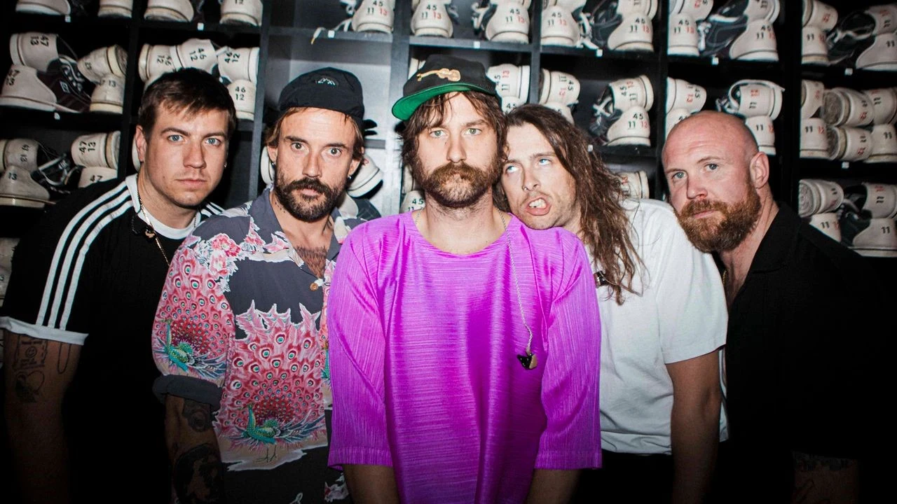 Idles wystąpią na Rock for People Pięciu mężczyzn pozujących razem na tle półek z równo ustawionymi białymi butami sportowymi, jeden z mężczyzn ubrany w różową bluzkę wyróżnia się na pierwszym planie.