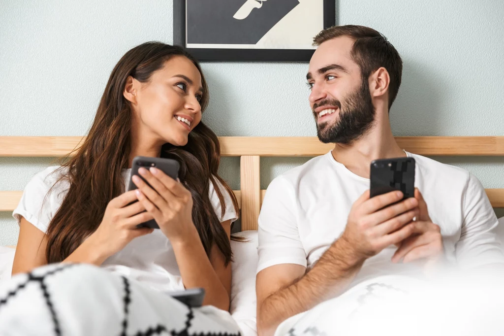 Telefon i nadmierne korzystanie z technologii może negatywnie wpłynąć na związek Młoda para siedzi razem na łóżku, patrząc na siebie z uśmiechem i trzymając smartfony w dłoniach, ubrani w białe koszulki, pod przykryciem.