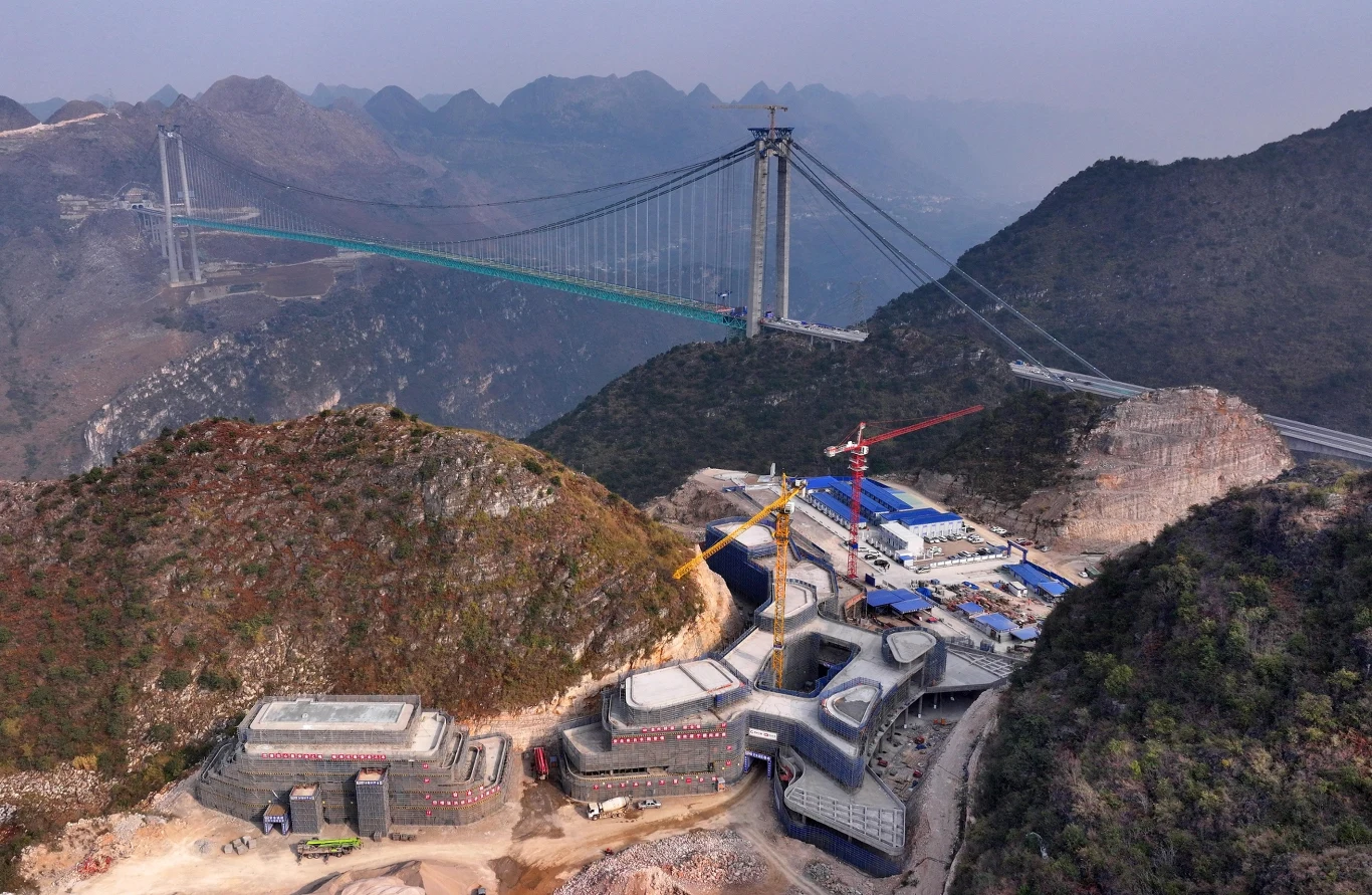 Huajiang Grand Canyon Bridge nie tylko umożliwi szybkie podróżowanie, ale i stanie się magnesem na turystów. Powstają tam centrum, kładki i platforma do skoków na bungee. Nowoczesny wiszący most rozpięty nad głęboką doliną w górskim krajobrazie, w tle wzniesienia pokryte roślinnością, w dolnej części kompleks budynków oraz maszyny budowlane, trwa rozbudowa infrastruktury.