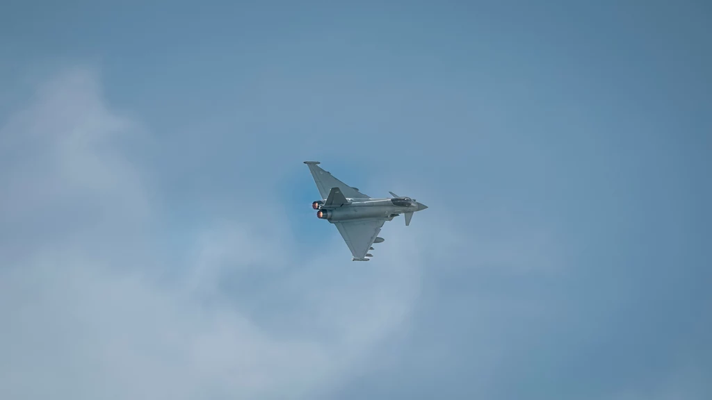 Nowoczesny myśliwiec wojskowy lecący na niebie, z widocznymi ciągami silnika i aerodynamiczną sylwetką na tle delikatnie zachmurzonego nieba.