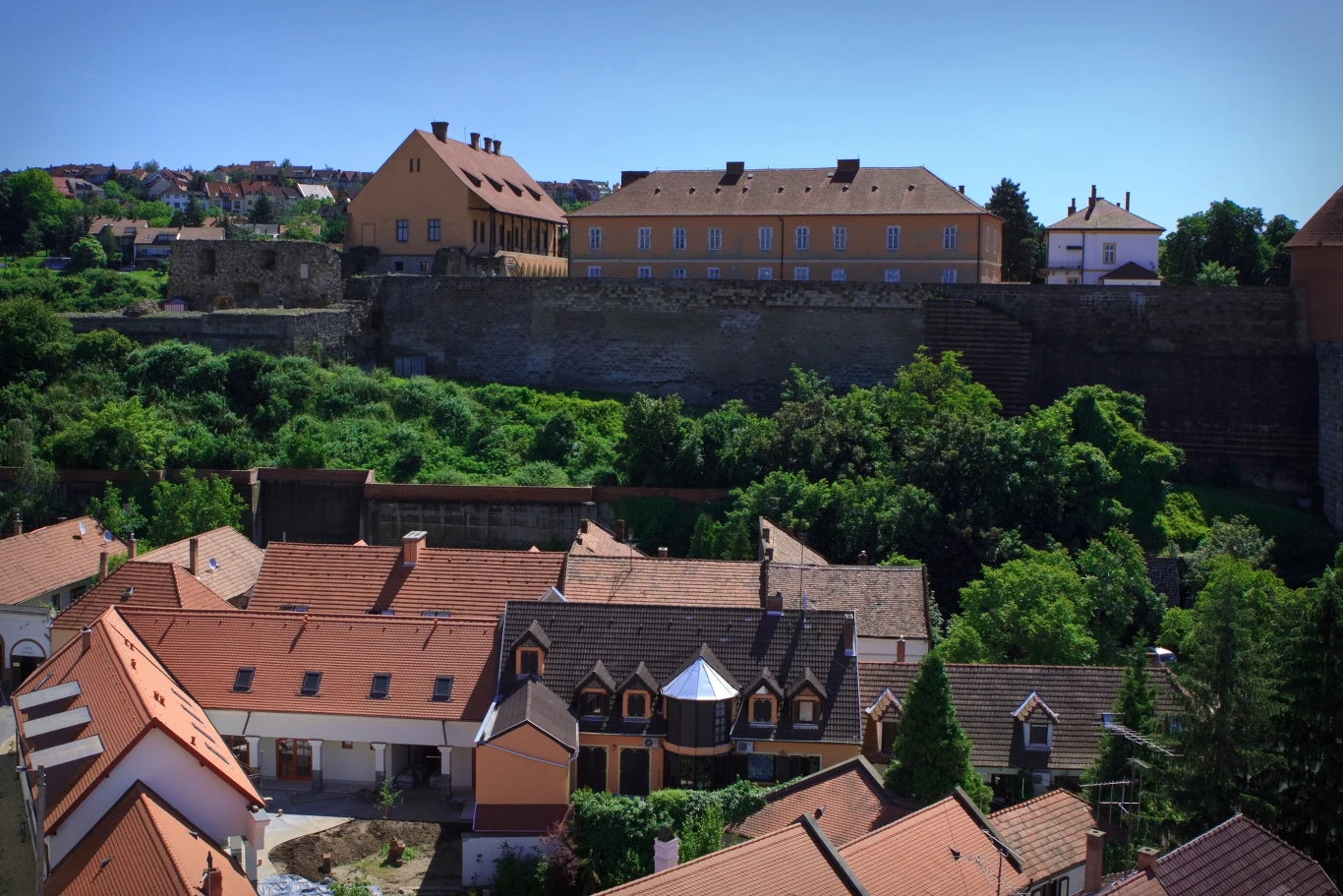 Widok na twierdzę Eger z minaretu Czerwone dachy domów w niskiej zabudowie, otoczone bujną zielenią, w tle wysoki mur obronny oraz duży historyczny budynek o ceglastej elewacji, z jasnoniebieskim niebem nad całością.