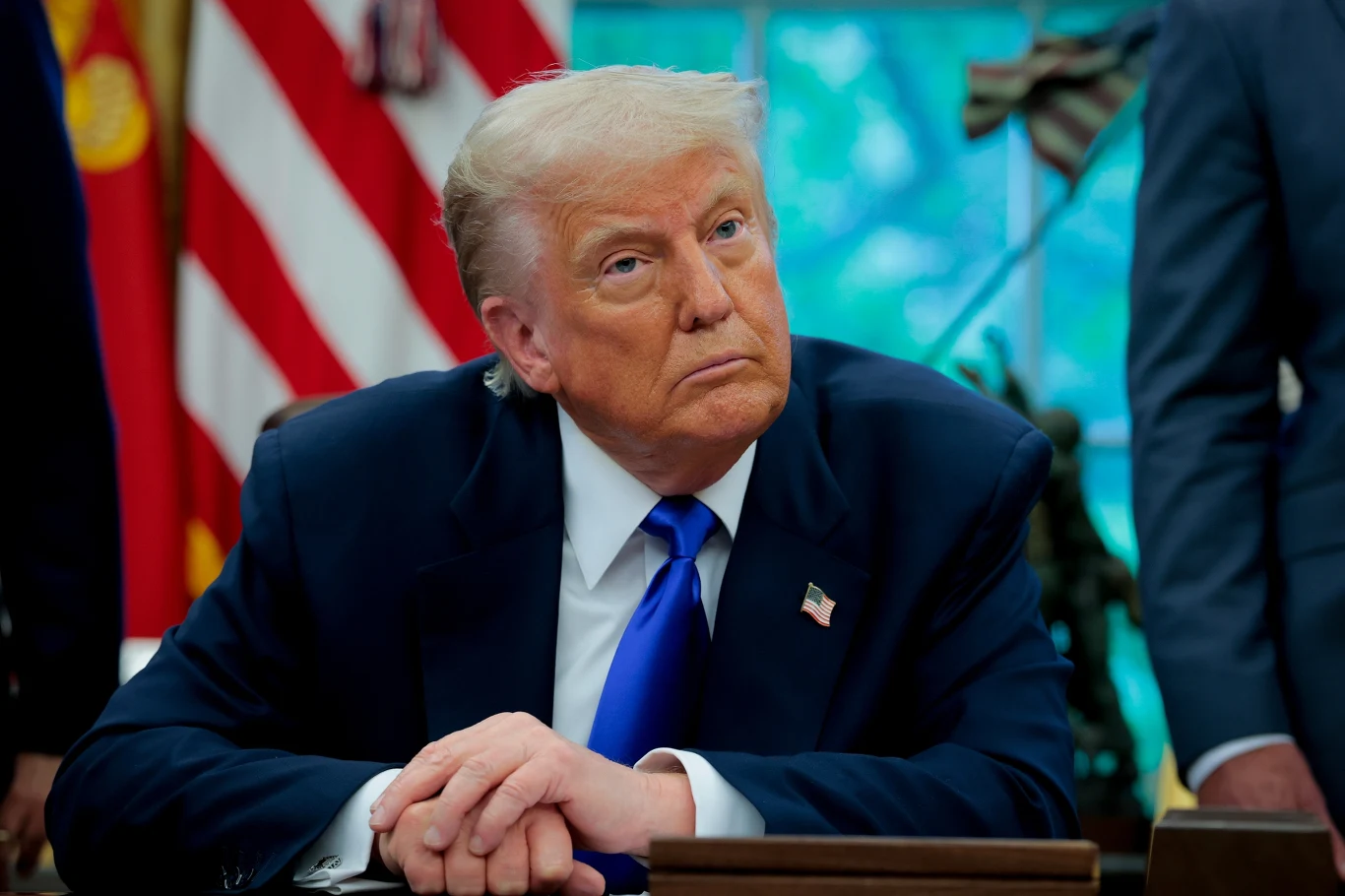 Korea Północna krytykuje plan Donalda Trumpa dotyczący budowy Złotej Kopuły nad USA Mężczyzna w garniturze z niebieskim krawatem siedzi za biurkiem, ma poważny wyraz twarzy, w tle widoczna flaga USA oraz fragment wnętrza urzędowego.