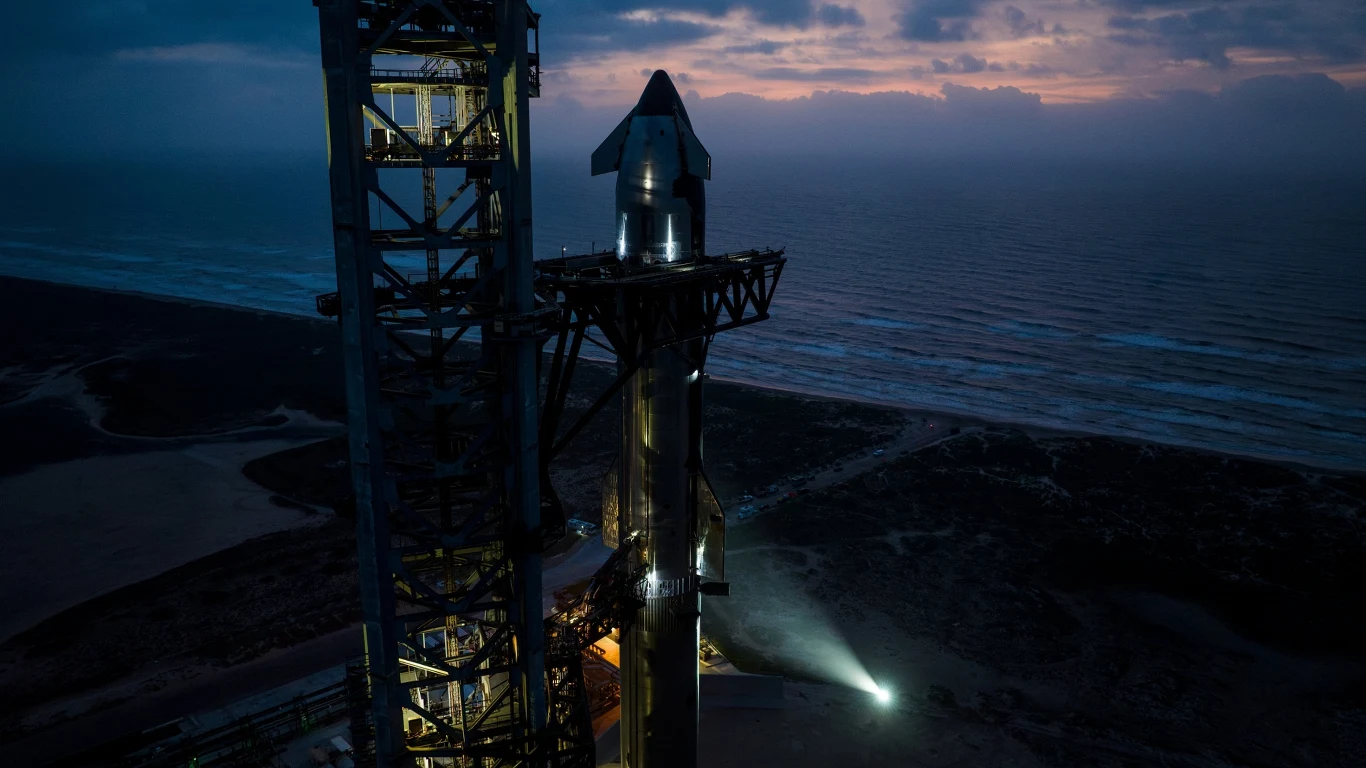 Starship gotowy do 9. lotu. SpaceX zrobi coś pierwszy raz. Rakieta przygotowana do startu na platformie, wokół metalowa konstrukcja i wieża startowa, w tle widok na ocean i niebo o zmierzchu