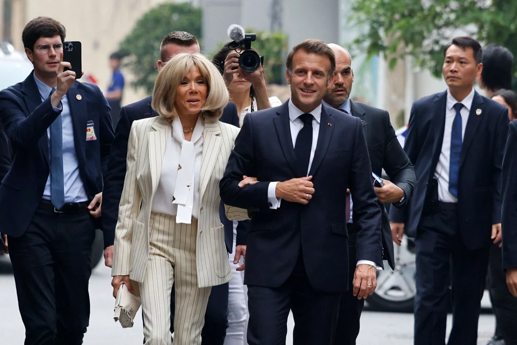 Brigitte Macron w eleganckim garniturze Emmanuel Macron w eleganckim granatowym garniturze idzie pod rękę z Brigitte Macron ubraną w jasny garnitur w prążki, otoczeni przez ochronę oraz osobę z aparatem i innych towarzyszących im ludzi, w tle widoczne miejskie otoczenie.