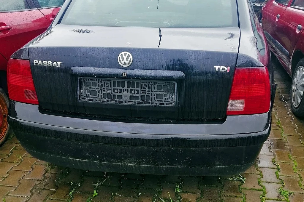 Czarny samochód marki Volkswagen Passat TDI zaparkowany na mokrym, brukowanym parkingu, brak tablic rejestracyjnych, widoczne ślady zabrudzeń na karoserii.