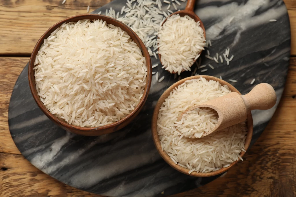 Dwie miski z nieugotowanym ryżem basmati ustawione na marmurowej desce, z dwoma drewnianymi łyżkami do nakładania ziaren, kilka ziaren rozsypanych wokół naczyń.