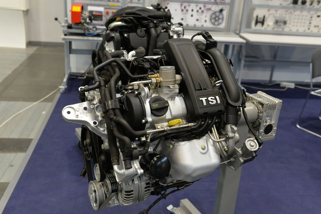 Silnik TSI podczas targów Poznań Motor Show 2023. Silnik samochodowy typu TSI z widocznymi przewodami, osprzętem i elementami układu zasilania, ustawiony na stojaku w pomieszczeniu laboratoryjnym.