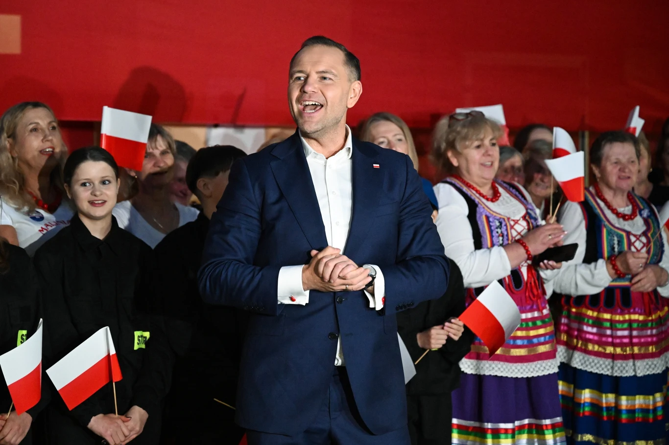 NIK skontrolował IPN. Instytucja, którą kierował Karol Nawrocki z szeregiem nieprawidłowości Mężczyzna w garniturze stoi na tle grupy ludzi, z których część ubrana jest w tradycyjne stroje ludowe, a wielu z nich trzyma biało-czerwone flagi Polski.