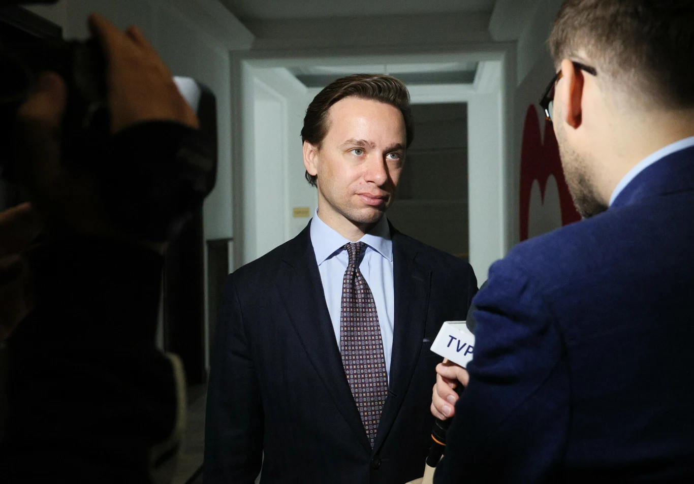 Krzysztof Bosak w garniturze udziela wywiadu dziennikarzowi TVP w jasno oświetlonym korytarzu, widoczny mikrofon i kamera. Krzysztof Bosak w garniturze udziela wywiadu dziennikarzowi TVP w jasno oświetlonym korytarzu, widoczny mikrofon i kamera.