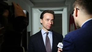 Krzysztof Bosak w garniturze udziela wywiadu dziennikarzowi TVP w jasno oświetlonym korytarzu, widoczny mikrofon i kamera.