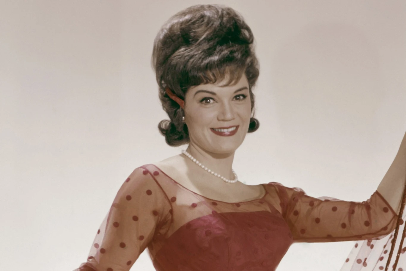 Connie Francis 63 lata temu stworzyła przebój "Pretty Little Baby" Elegancko ubrana kobieta w bordowej sukni z przezroczystymi rękawami i groszkami, z krótkim naszyjnikiem z pereł, uśmiecha się na jasnym tle, włosy zaczesane w stylową fryzurę z lat 60.