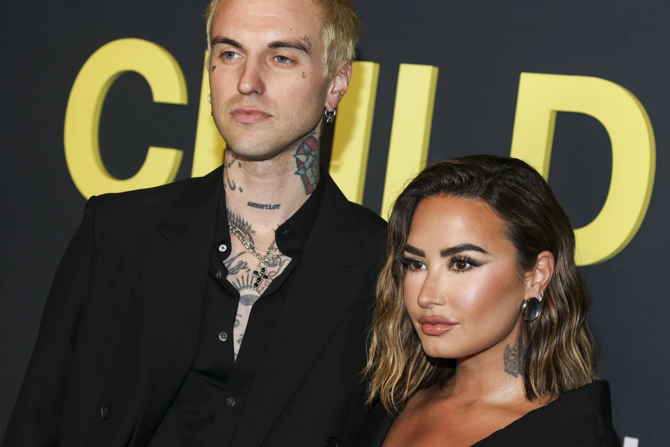 Demi Lovato wyszła za mąż za Jordana Lutesa Dwoje elegancko ubranych ludzi pozuje razem na tle ciemnej ściany z dużymi żółtymi literami, mężczyzna z licznymi tatuażami na szyi i klatce piersiowej wraz z kobietą o profesjonalnym makijażu i krótkich, falowanych włosach.