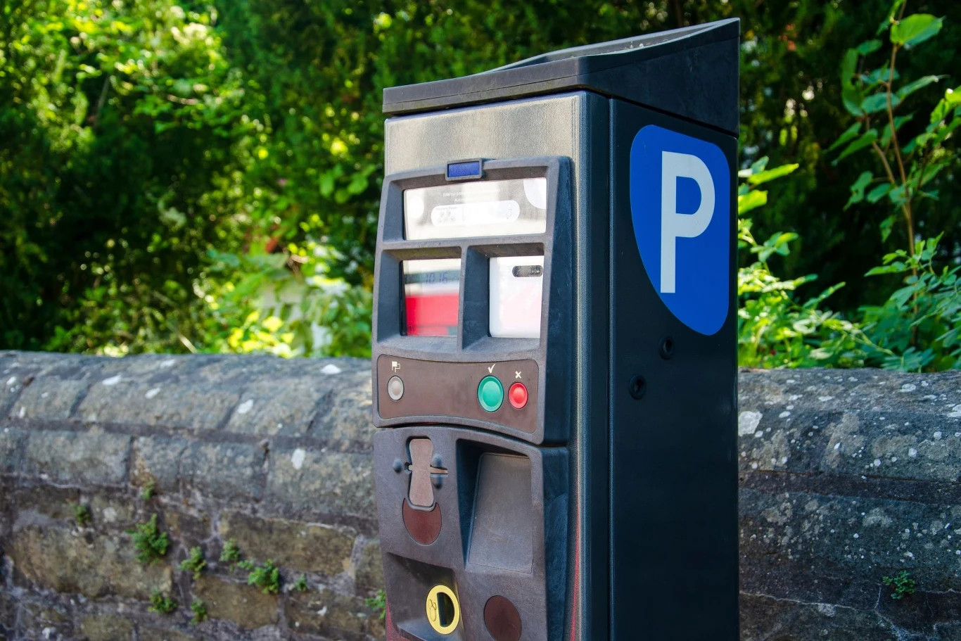 Osoby płacące przez parkometr mają czas na opłatę, inaczej niż użytkownicy aplikacji mobilnych. Automat parkingowy umieszczony przy kamiennym murze, otoczony roślinnością, z niebieskim oznaczeniem 'P' sugerującym miejsce parkingowe.