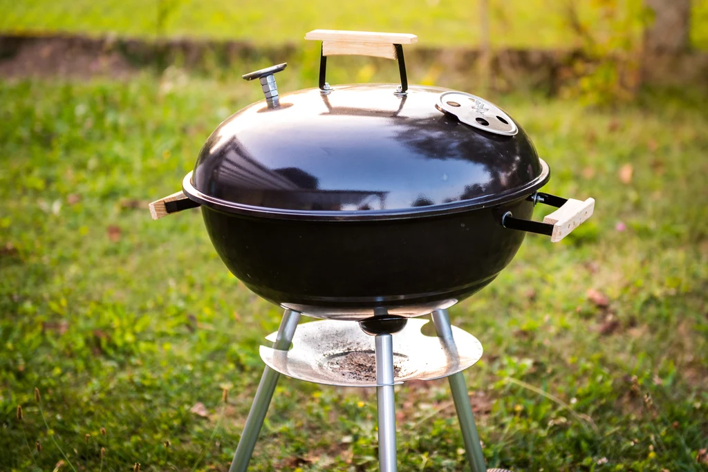 Staraj się ustawić ruszt stosunkowo wysoko nad źródłem ciepła Metalowy grill ogrodowy z kopułową pokrywą i drewnianymi uchwytami stoi na zielonej trawie w słoneczny dzień.