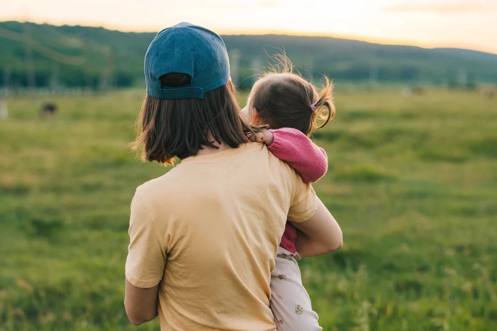 Wiele macoch uznało określenie "bonusowa mama" za bliższe ich sercom Osoba w jasnej koszulce i niebieskiej czapce trzyma na rękach małe dziecko w różowej bluzie, stojąc tyłem do aparatu na tle zielonej łąki i wzgórz, przy delikatnym świetle zachodzącego słońca.
