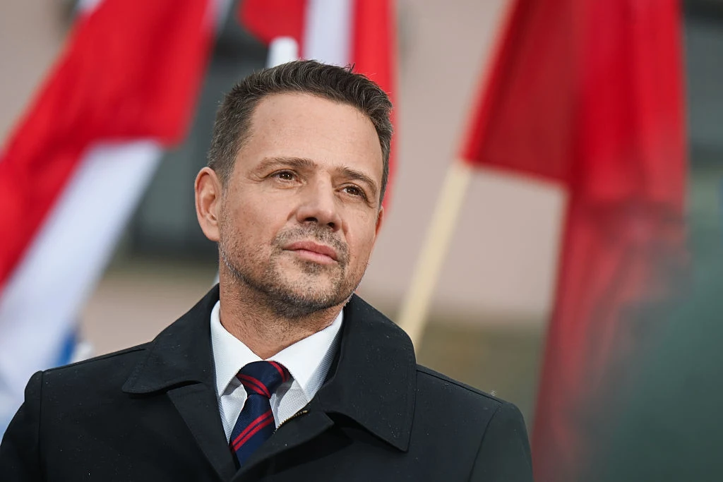 Mężczyzna w eleganckim płaszczu i krawacie stoi na tle rozmytych czerwonych i biało-czerwonych flag, patrząc przed siebie z neutralnym wyrazem twarzy.