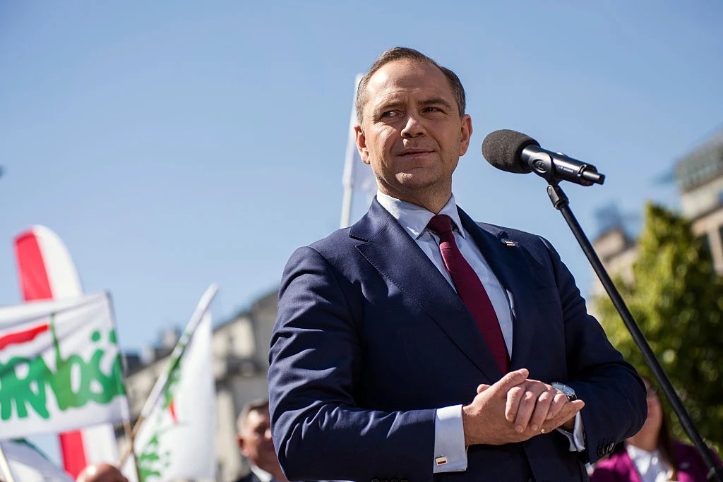 Mężczyzna w garniturze i czerwonym krawacie stoi na zewnątrz przy mikrofonie, w tle widoczne flagi z napisem 'Solidarność' i uczestnicy zgromadzenia, słoneczna pogoda.