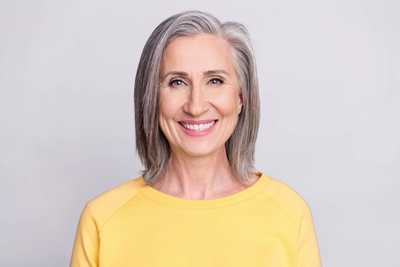Doświadczona, uśmiechnięta kobieta z siwymi włosami w żółtej bluzie na jasnym tle, wyrażająca radość i pozytywną energię.
