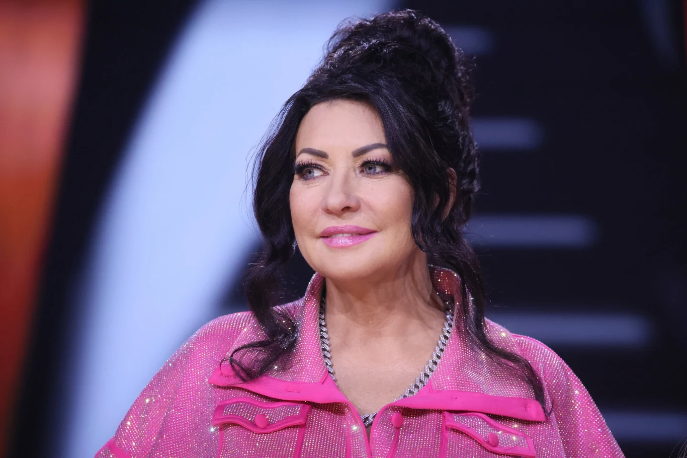 Alicja Węgorzewska to jedna z najsłynniejszych polskich śpiewaczek operowych Kobieta o ciemnych włosach spiętych w kok, ubrana w różową, błyszczącą kurtkę, z delikatnym makijażem, stoi na tle rozmytego czarno-białego wzoru.