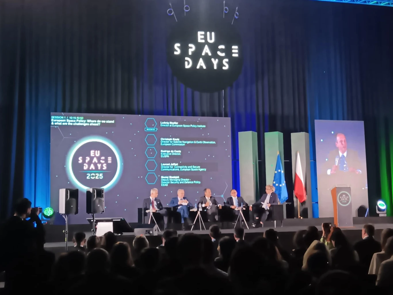 Panel dyskusyjny podczas konferencji EU Space Days 2025, na scenie pięciu prelegentów siedzących przy stolikach, duży ekran z agendą wydarzenia, po prawej stronie mównica i przemawiający mężczyzna, w tle flagi Unii Europejskiej i Polski, widownia w pół...