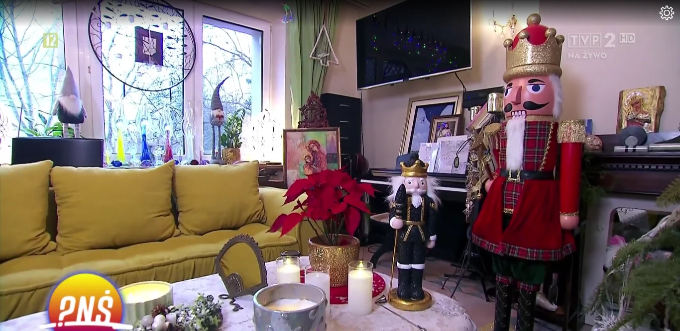 Tak wygląda salon Alicji Węgorzewskiej Wnętrze udekorowane na święta, z żółtą kanapą, choinką, dużą figurą dziadka do orzechów w czerwonym stroju, świecami na stoliku, rośliną gwiazdą betlejemską oraz obrazami i dekoracjami świątecznymi wokół pianina.