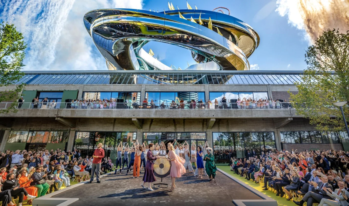 Gdy historia spotyka nowoczesność. Fenix to nowy symbol Rotterdamu Nowoczesny budynek o futurystycznych kształtach wznosi się ponad zgromadzeniem ludzi, którzy świętują wydarzenie na zewnątrz; grupa elegancko ubranych osób stoi przy dużym gongu, w tle liczna publiczność oraz szklana fasada.