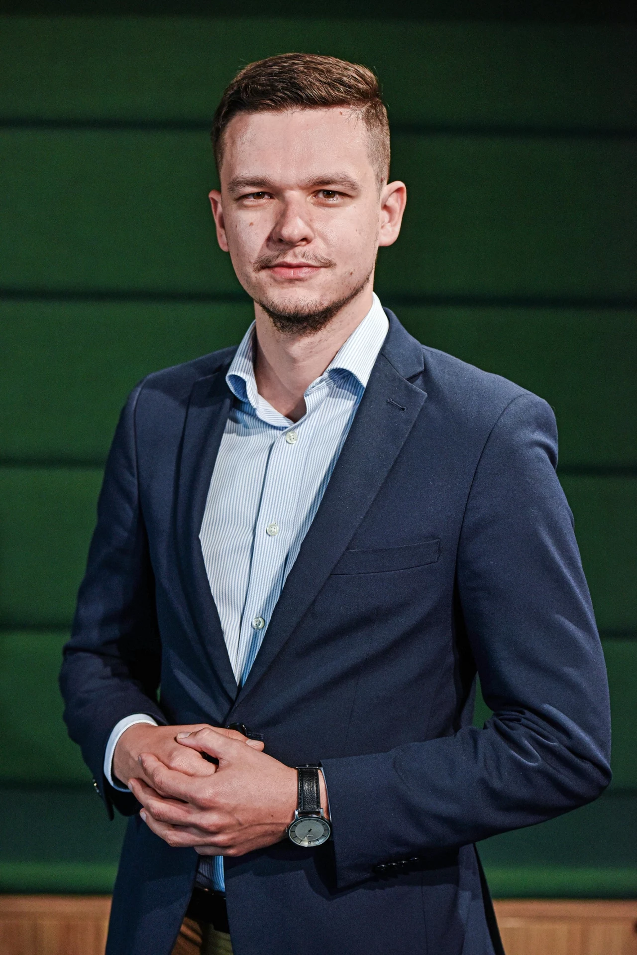 Młody mężczyzna w granatowym garniturze i jasnej koszuli stojący na tle zielonych paneli ściennych, z poważnym wyrazem twarzy, trzymający dłonie splecione przed sobą.