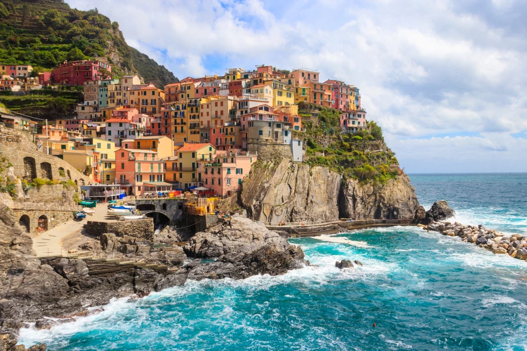 Miasteczko Manarola stało się wizytówką Cinque Terre Kolorowe domy osadzone na stromym klifie u wybrzeża morza, fale rozbijające się o skały, błękitna woda oraz część portu z zacumowanymi łodziami, górzyste tło i niebo z chmurami.