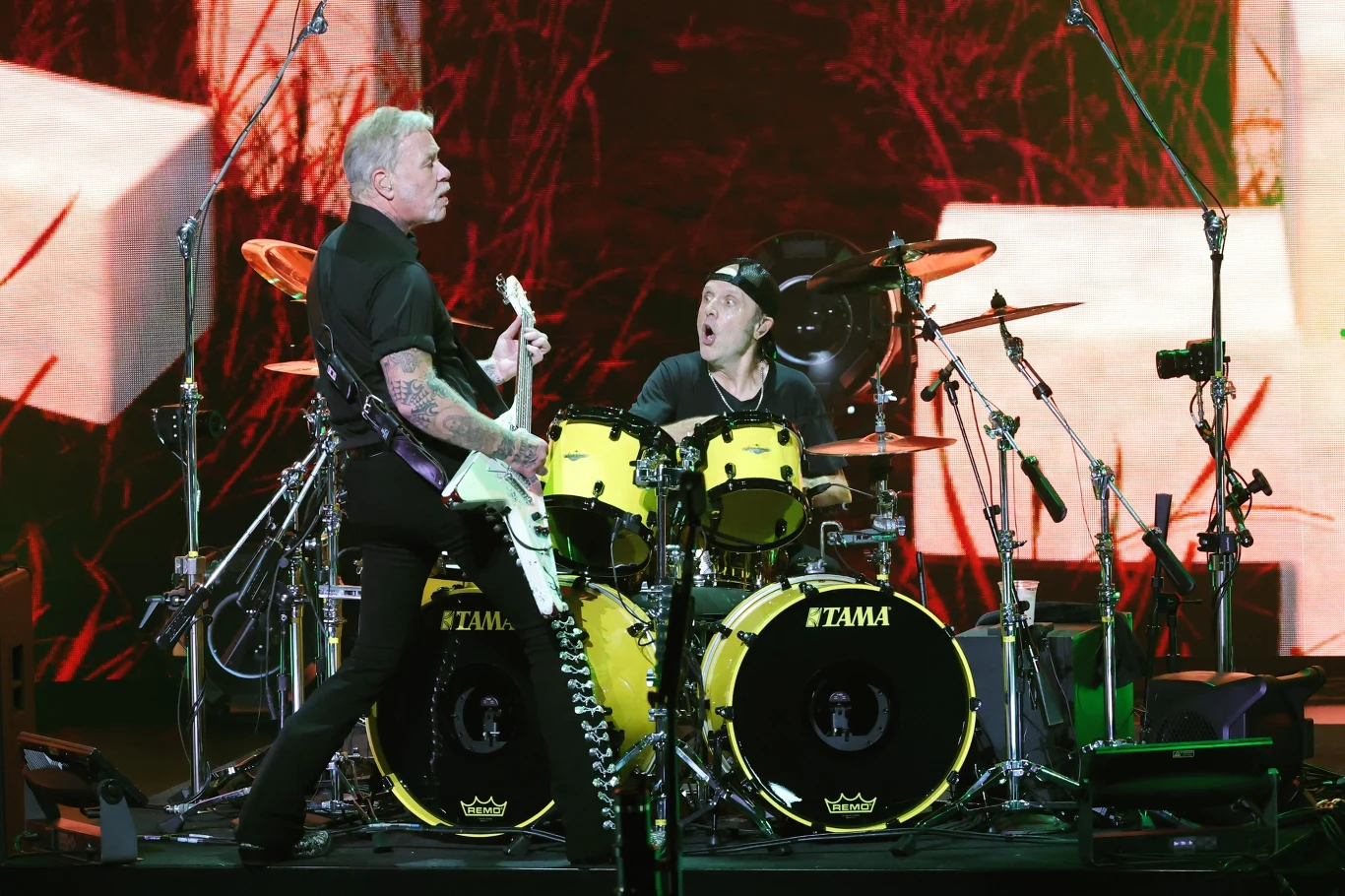 James Hetfield i Lars Ulrich (Metallica) w akcji Dwóch muzyków rockowych podczas koncertu, jeden gra na gitarze elektrycznej, drugi na żółtym zestawie perkusyjnym, intensywne oświetlenie sceniczne oraz tło z dużymi ekranami, dynamiczna atmosfera występu.