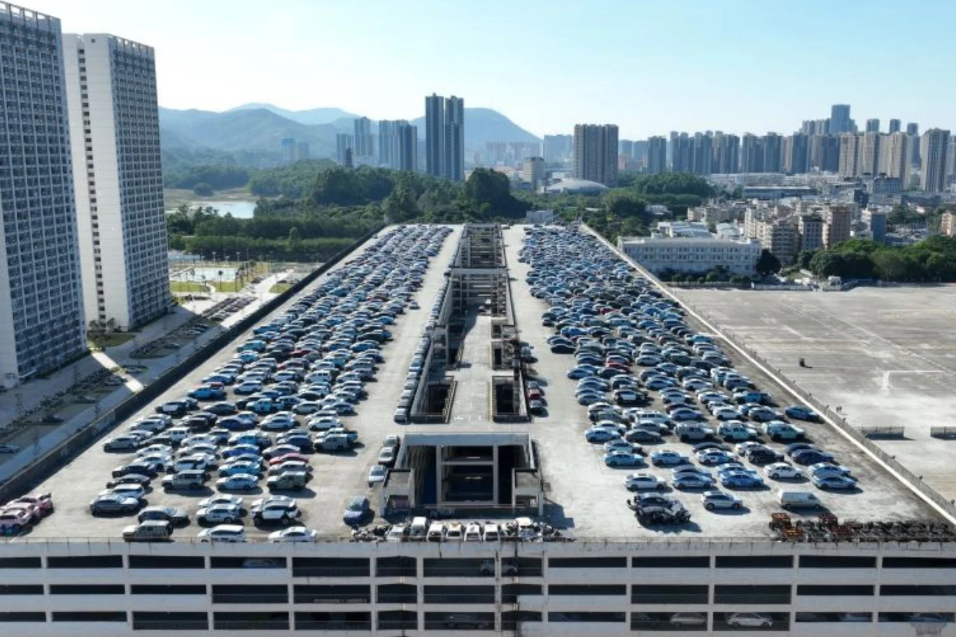 Rooftop dużego parkingu z setkami zaparkowanych samochodów, otoczony wysokimi budynkami mieszkalnymi i panoramą miasta w tle, jasne niebo i widoczne odległe wzgórza przyczyniają się do miejskiego charakteru scenerii.