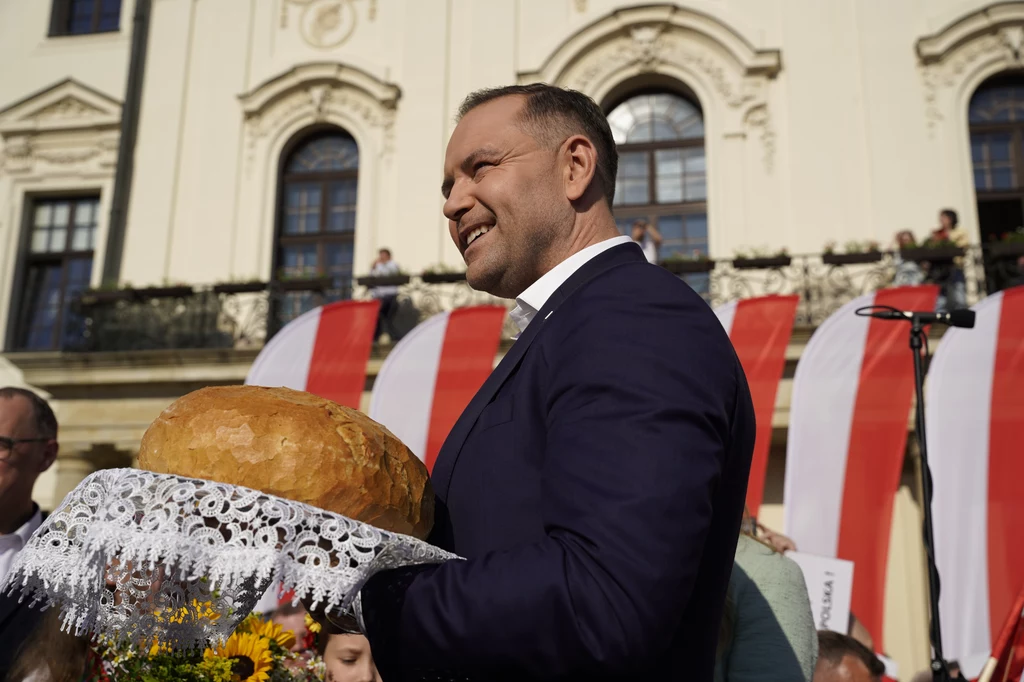Mężczyzna w garniturze trzyma na eleganckiej serwecie okrągły bochenek chleba podczas uroczystości plenerowej z biało-czerwonymi dekoracjami na tle historycznego budynku