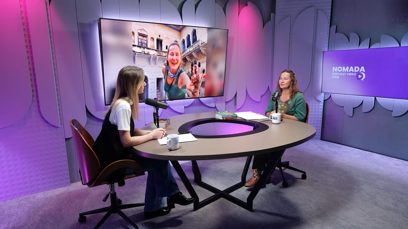 Podcast Nomada: Rozmowa z Kingą Lityńską o trudach życia w Indiach Dwie kobiety siedzą przy owalnym stole w profesjonalnym studiu podcastowym, rozmawiając przy mikrofonach, na ścianie za nimi widoczny jest ekran z dużym zdjęciem uśmiechniętej kobiety na tle budynków.