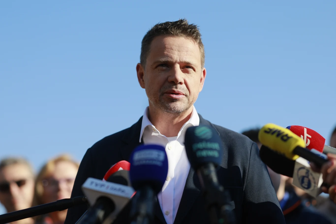 Rafał Trzaskowski w trakcie kampanii wyborczej Mężczyzna w garniturze stoi przed mikrofonami kilku różnych stacji telewizyjnych, udzielając wypowiedzi mediom na tle nieba, w otoczeniu ludzi.