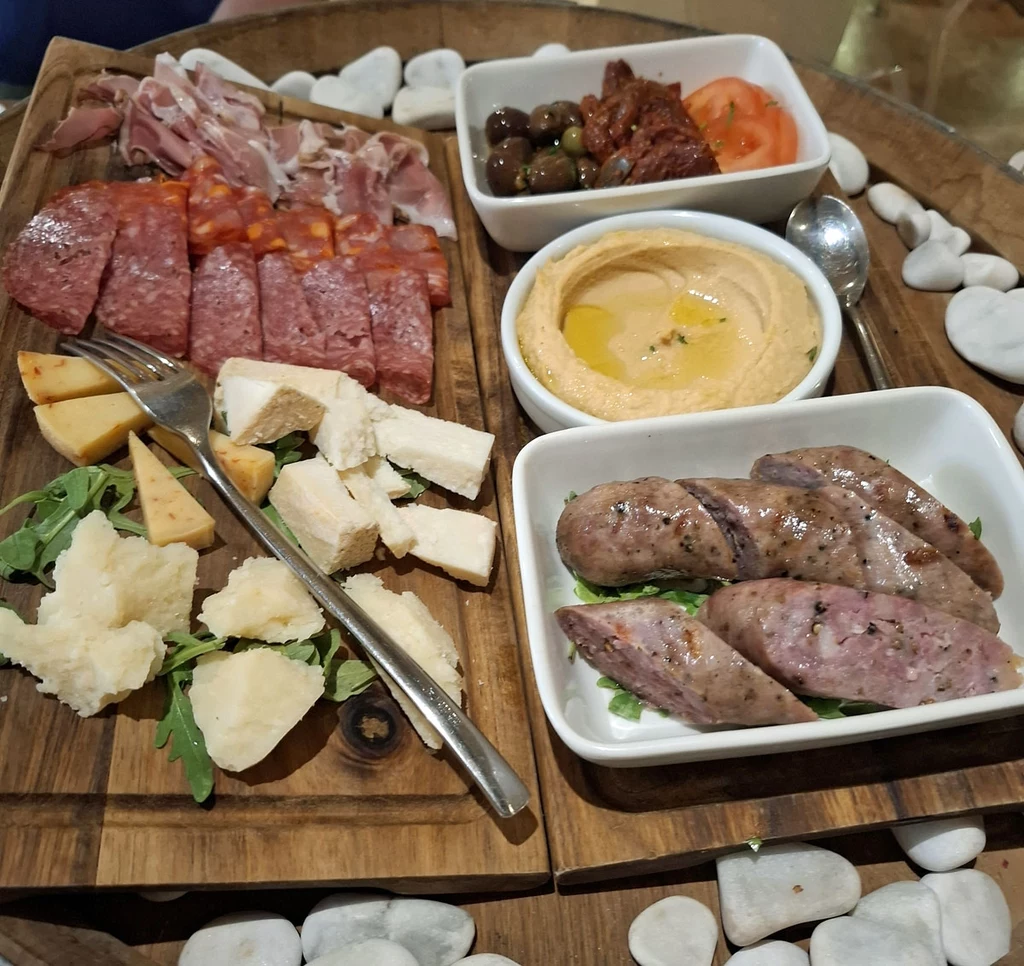 Vini e Capricci na Gozo. Owcze sery, maltańska kiełbasa, humus i oliwki to popularne przekąski. Deska z różnymi rodzajami wędlin, serem, kawałkami kiełbasy oraz dodatkami takimi jak hummus, oliwki, suszone pomidory i rukola, wszystko podane na drewnianej desce dekorowanej białymi kamieniami.