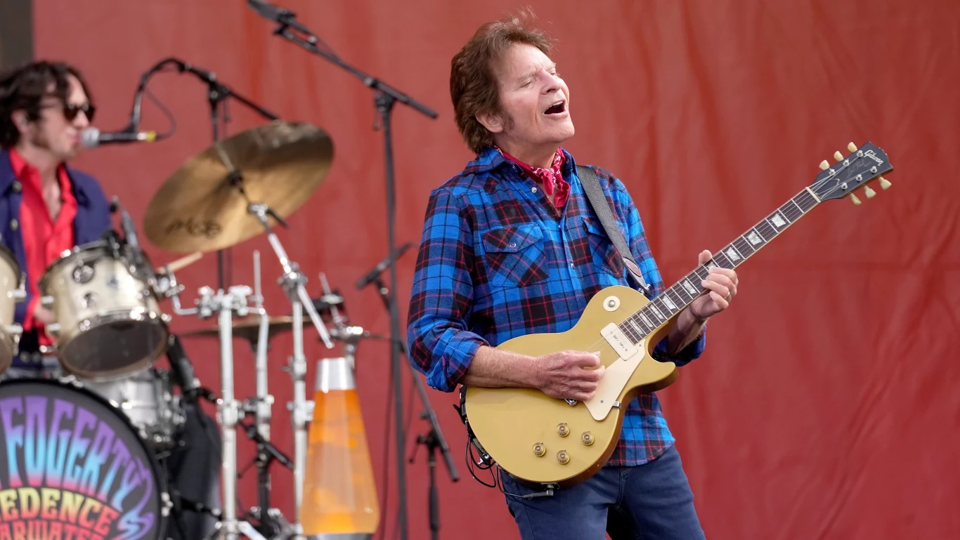 John Fogerty był liderem Creedence Clearwater Revival Mężczyzna w niebieskiej koszuli w kratę gra na gitarze elektrycznej na scenie podczas występu na żywo, w tle perkusista oraz sprzęt muzyczny, całość przed czerwonym tłem.