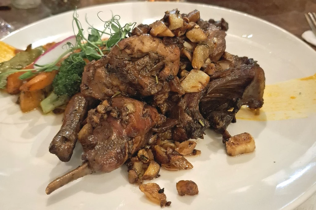 Królik duszony w restauracji United Danie składające się z duszonego mięsa królika z kawałkami czosnku, podane z gotowanymi warzywami, ozdobione delikatnymi kiełkami na białym talerzu.