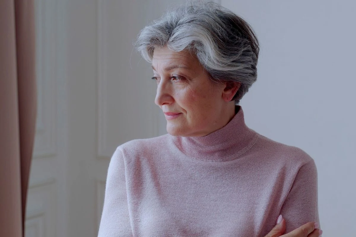 Starsza kobieta o siwych włosach w różowym golfie, stojąca z założonymi ramionami i patrząca spokojnie w bok. Minimalistyczne jasne tło podkreśla wyciszony charakter ujęcia.