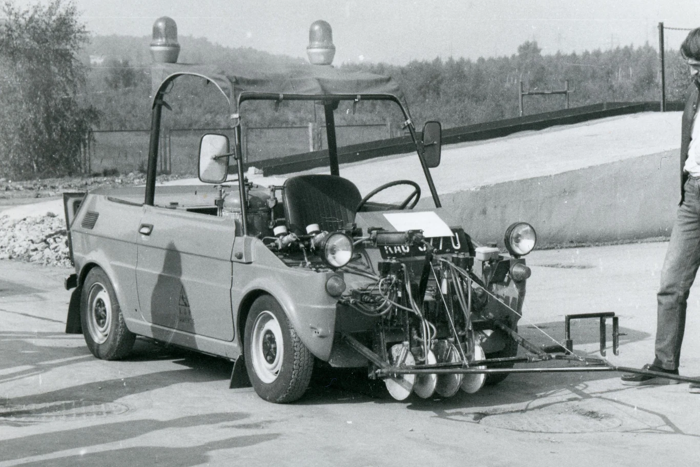 Pingwin 04 w pełnej okazałości. Powstał na bazie popularnego Fiata 126p. Mały pojazd testowy z otwartą kabiną wyposażony w liczne urządzenia techniczne umieszczone z przodu, takie jak lampy, przewody i czujniki, z przystawką pomiarową połączoną z przodem auta, a obok pojazdu stoi człowiek; w tle zalesiony teren oraz infrast...