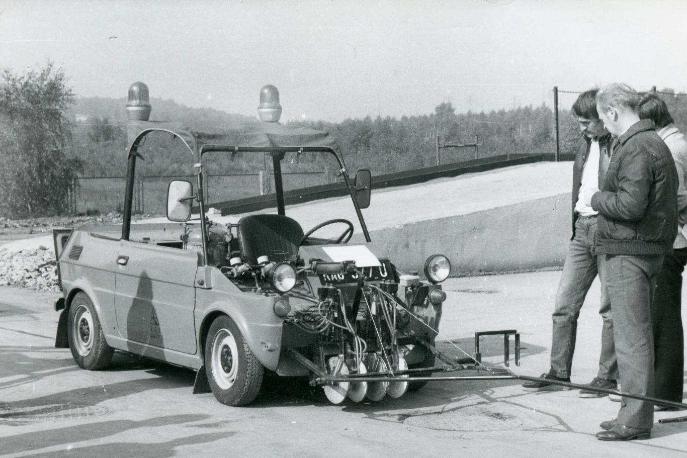 Kierowca-operator obsługiwał urządzenie malujące z wnętrza kabiny. Prędkość robocza wynosiła 7 km/h. Mały samochód wyposażony w aparat pomiarowy lub eksperymentalne urządzenia techniczne, wokół pojazdu stoi kilku mężczyzn obserwujących pojazd. Tło stanowi asfaltowa droga oraz lekko zalesiony teren.