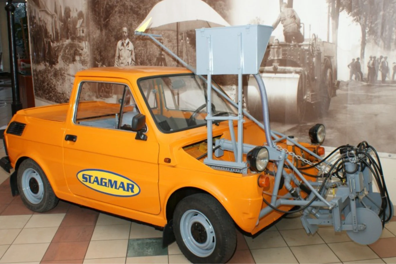 Pingwin 04 o numerze seryjnym 011. Ten egzemplarz kosztował 5,1 mln zł. Pomarańczowy samochód marki Stagmar wyposażony w specjalistyczną maszynę montowaną na jego przedzie, służącą prawdopodobnie do prac drogowych lub znakowania nawierzchni, stojący wewnątrz budynku o kafelkowanej podłodze.