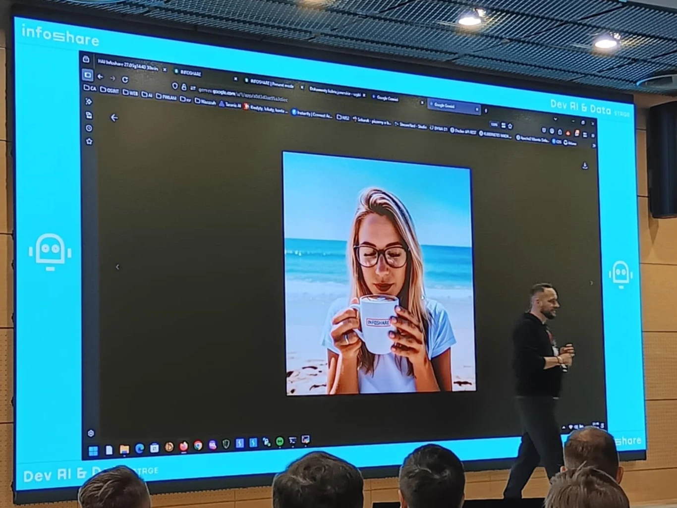 Duży ekran prezentacyjny z wyświetlonym zdjęciem młodej kobiety pijącej kawę na tle plaży, z boku stoi mężczyzna trzymający mikrofon i prowadzący prezentację, na widowni widoczne sylwetki kilku osób.