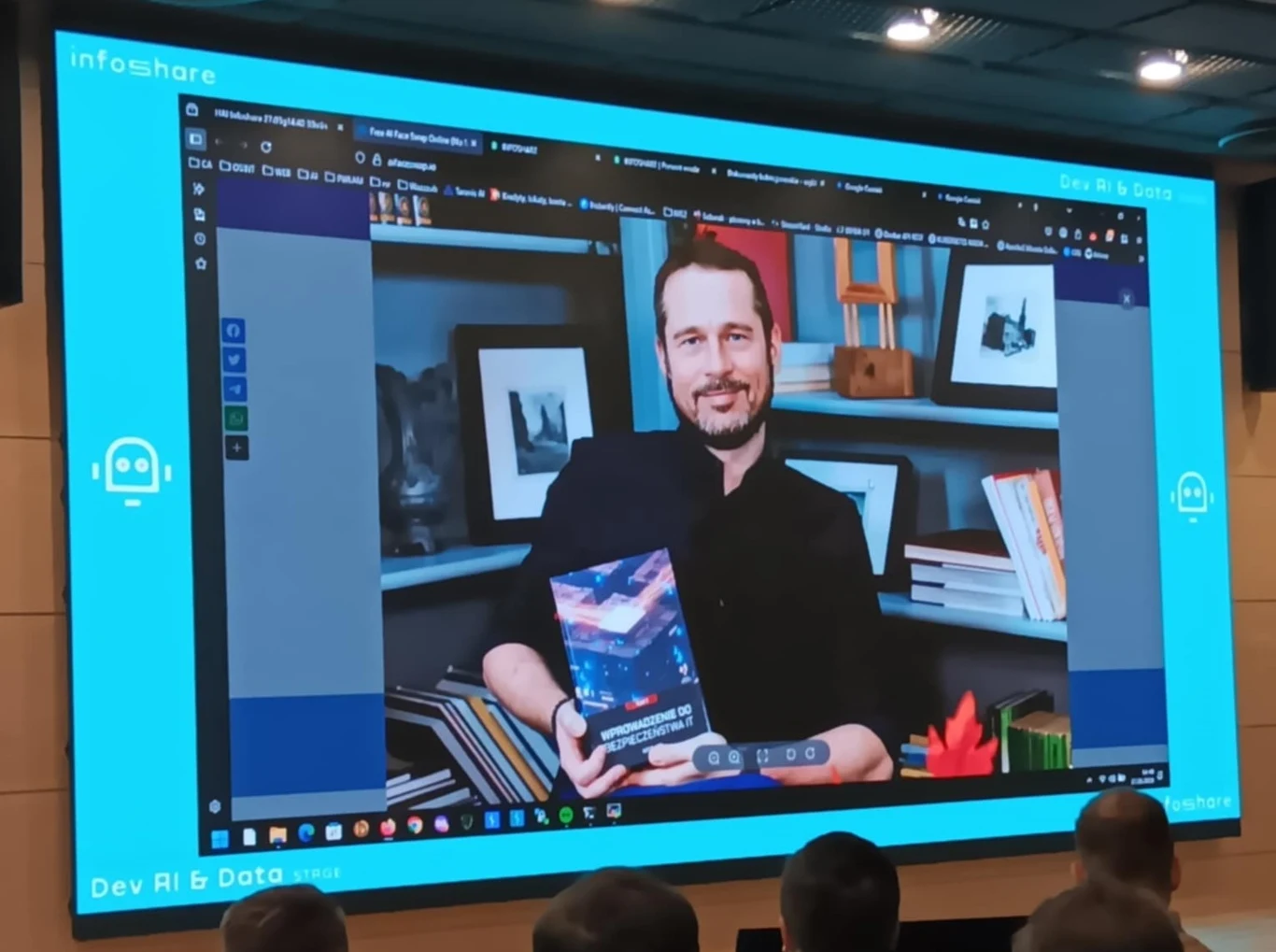 Mężczyzna trzymający książkę siedzi przy biurku na tle półek z książkami, jego zdjęcie jest wyświetlane na dużym ekranie podczas konferencji; na sali widoczni są uczestnicy obserwujący prezentację.