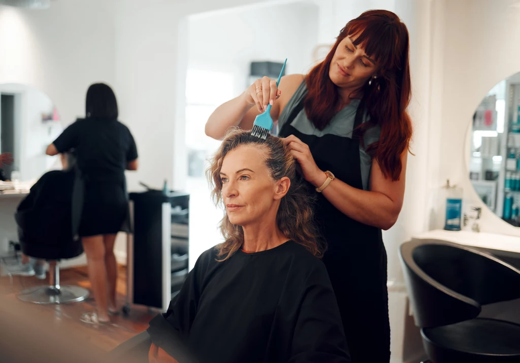 Balayage i refleksy odmieniają włosy dojrzałe, dodając im blasku i objętości Fryzjerka nakłada farbę na włosy dojrzałej kobiety siedzącej na fotelu w salonie fryzjerskim, w tle widoczna inna klientka oraz elementy wyposażenia salonu.