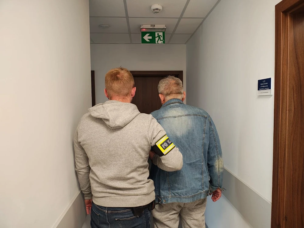 Dwóch mężczyzn idzie korytarzem, jeden trzyma drugiego za ramię, mają na sobie casualowe ubrania, jeden z nich ma opaskę na ramieniu z napisem 'POLICJA', drzwi i tabliczka na ścianie sugerują przestrzeń urzędową.