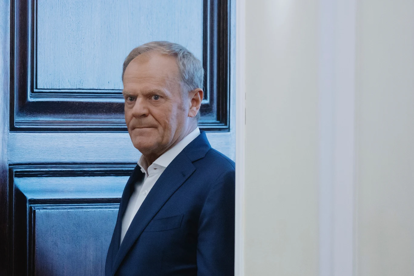 Donald Tusk i jego niebezpieczna kampania Mężczyzna w garniturze patrzący poważnym wzrokiem, stojący w uchylonych drzwiach, w tle ciemne drewniane drzwi kontrastujące z jasną ścianą.